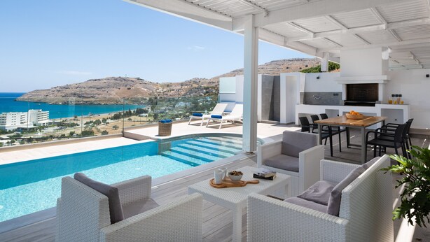 A modern outdoor living area with a pool, white wicker furniture, and a view of the sea and hills. - Villa Faros (Rhodes)