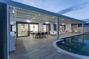 A modern outdoor pool area with a covered patio, a dining area, and a kitchen visible through glass doors. - Villa Faros (Rhodes)