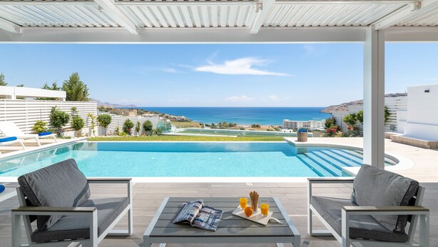 A poolside area with two chairs, a table with a magazine and drinks, and a view of the sea and landscape. - Villa Vrachos (Rhodes)