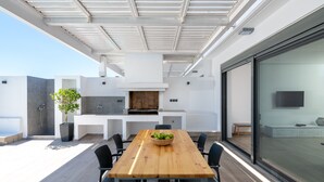 A modern outdoor dining area with a wooden table and chairs, a grill, and a large window. - Villa Vrachos (Rhodes)
