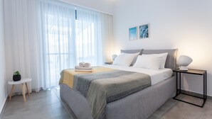 Une chambre à coucher bien rangée, avec un lit, des tables de chevet, une petite table d’appoint et une fenêtre ornée de rideaux légers.