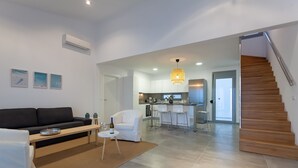 Un salon moderne avec un canapé noir, une petite table basse et un fauteuil blanc. On aperçoit à l’arrière-plan un coin cuisine avec un bar et un escalier.