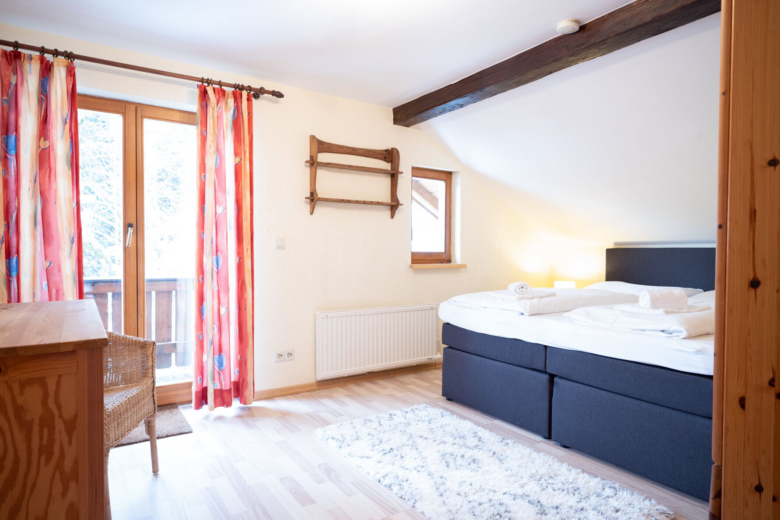 Une chambre à coucher comprenant un lit, une commode en bois, une chaise en osier, un radiateur et une fenêtre avec des rideaux.