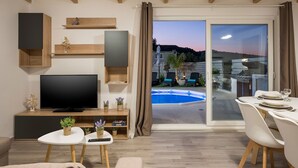 A modern living room with a flat-screen TV, wooden shelves, and a poolside dining area with a table set for two. - B&M Villa Sirili (Platanias)