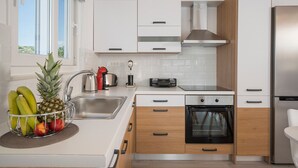 A modern kitchen with white cabinets, a stainless steel refrigerator, a built-in oven, and a fruit basket on the counter. - B&M Villa Sirili (Platanias)