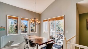 A dining area with a wooden table, chairs, a chandelier, and large windows with blinds.