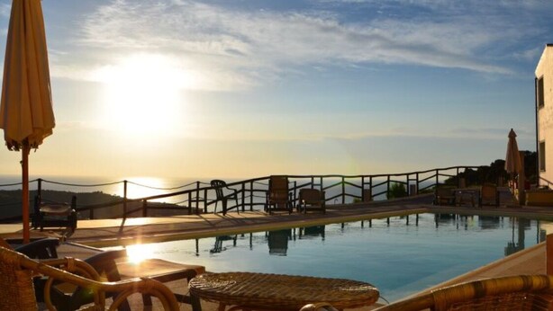 Un área de piscina con sillones de descanso y sombrillas, una valla de madera y vista al mar durante el atardecer.
