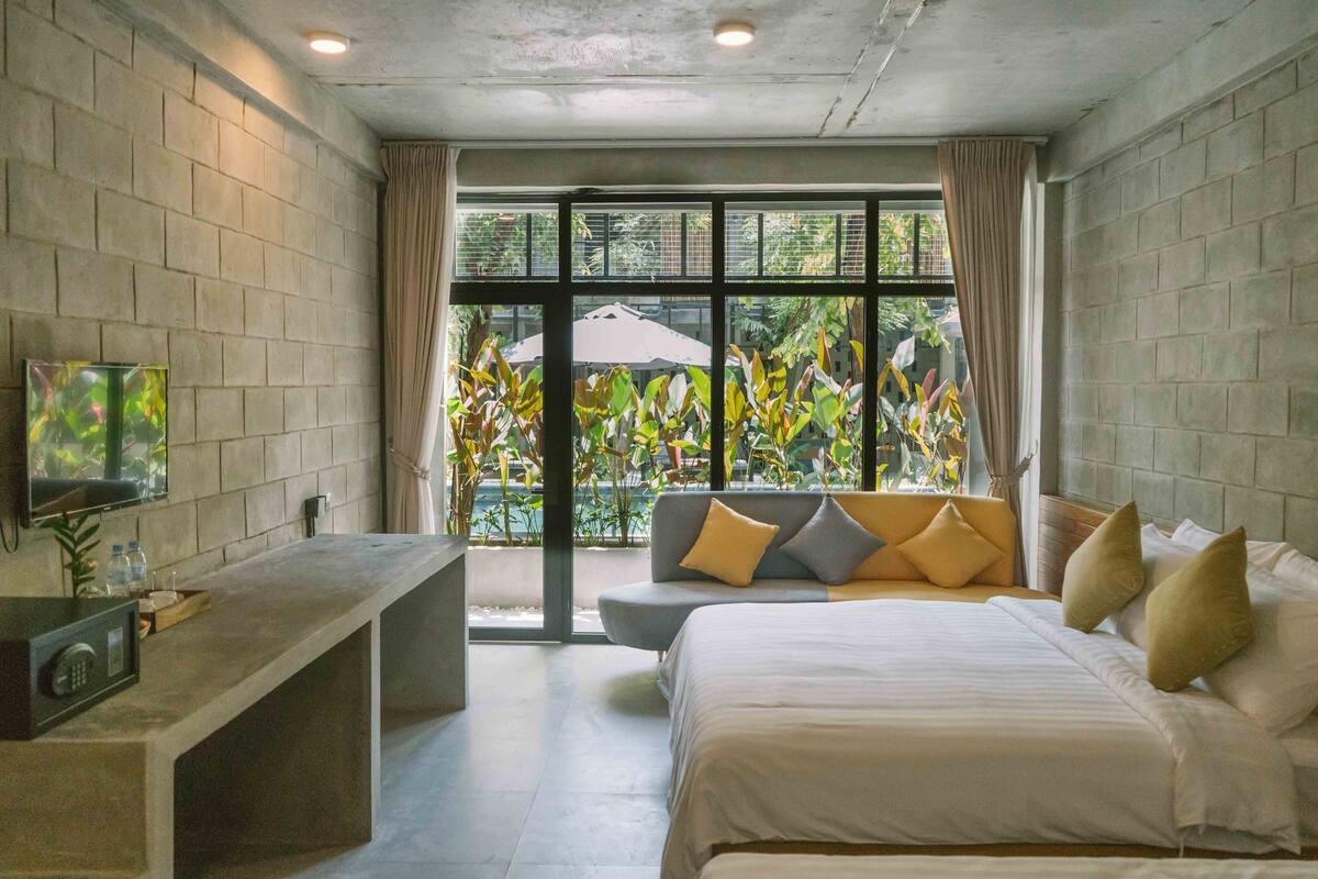A modern hotel room with a large bed, a seating area with cushions, a television, and a view of greenery outside.