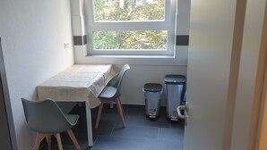 A small, clean room with a table and chairs, a window with a view of trees, and two trash bins. - Privatzimmer EKO 1 (Kassel)