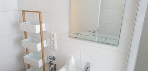 A modern bathroom with white tiles, a glass shelf, a wooden rack, a mirror, and a sink. - Privatzimmer EKO 1 (Kassel)