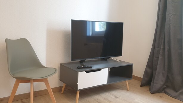 A modern living room with a flat-screen TV on a stand, a grey chair, and a white cabinet. - Privatzimmer EKO 1 (Kassel)
