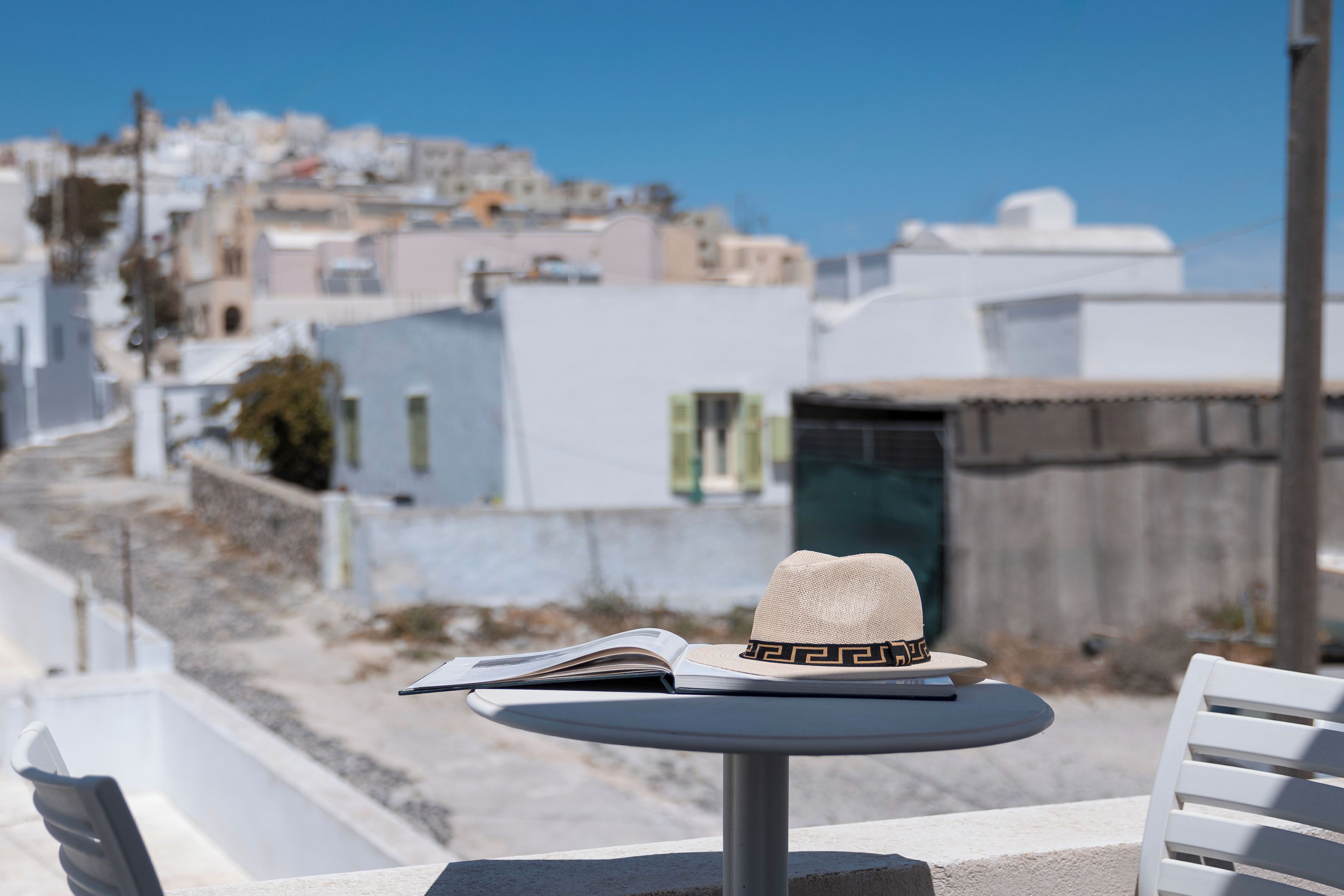 A table with a hat and book, overlooking a coastal town with white buildings and a clear sky.