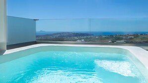A modern infinity pool with clear blue water, surrounded by a glass balustrade, offering a panoramic view of a coastal landscape and distant mountains. - Cinnamon Villa with Private Hot Tub (Santorini)
