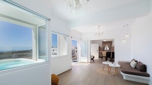 A modern living room with a large window, a sofa, a dining area, and a view of the ocean. - Cinnamon Villa with Private Hot Tub (Santorini)