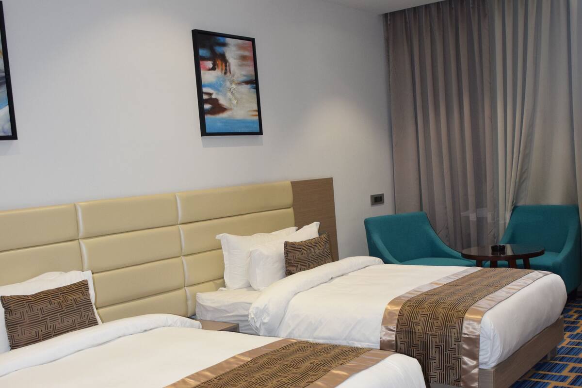 A hotel room with two beds, a headboard, a nightstand, a chair, a table, and a large window with curtains.
