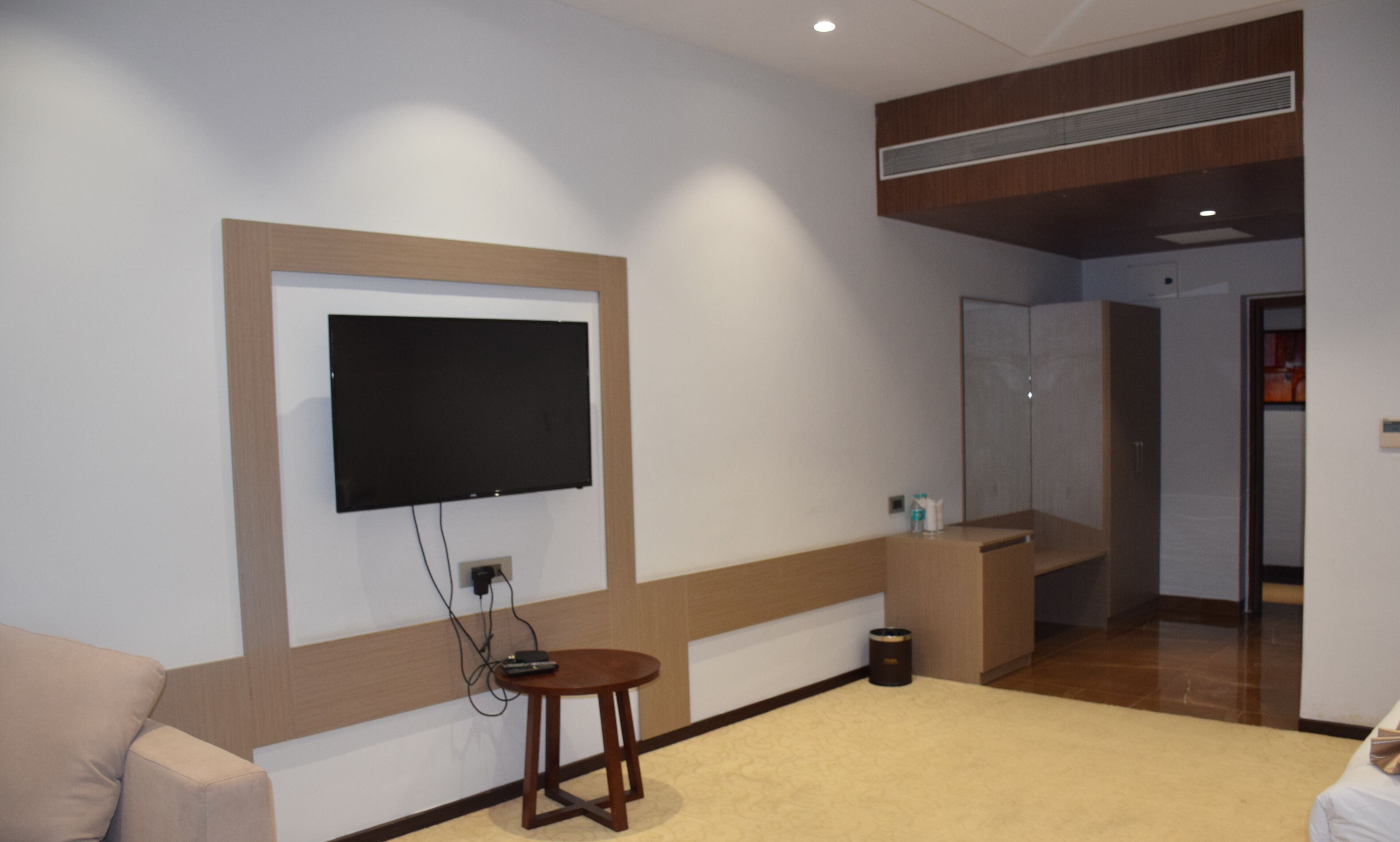 A modern hotel room with a flat-screen TV mounted on the wall, a small wooden table, a beige carpet, and a wooden accent wall.