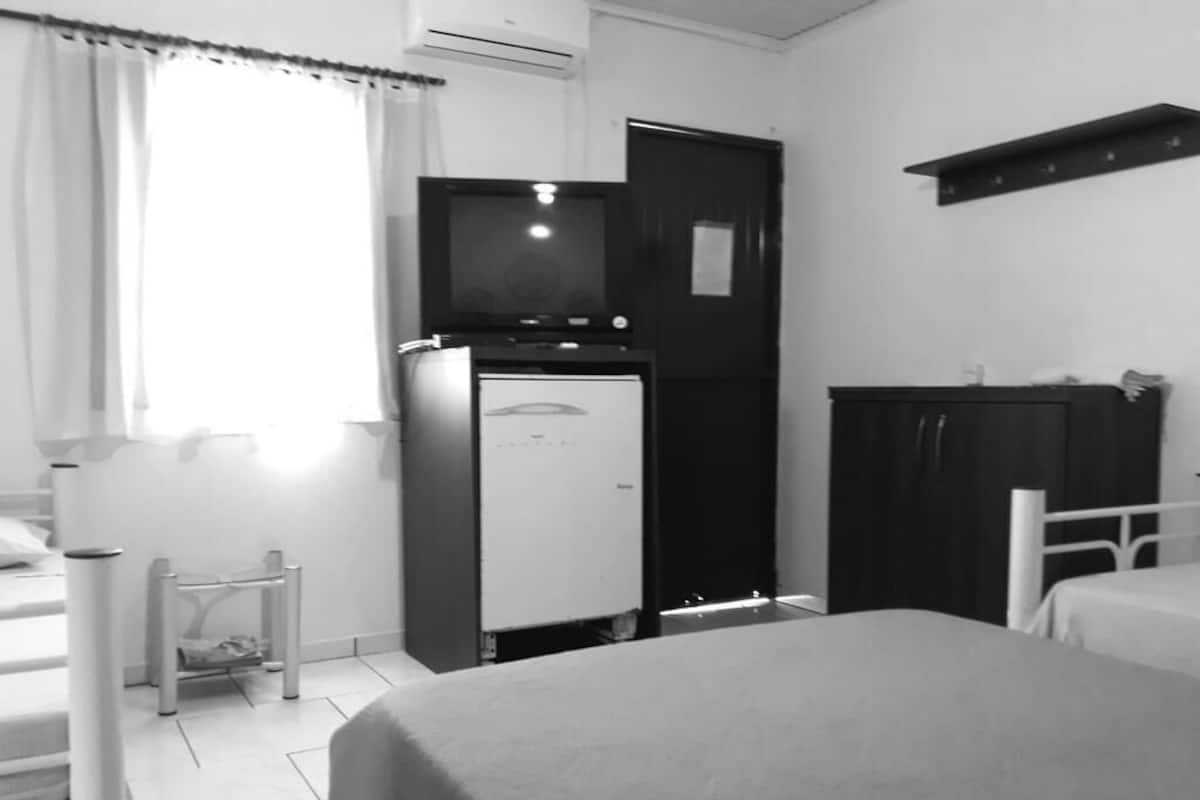 A compact hotel room with a bed, a small table, a wardrobe, a kitchenette with a refrigerator, and a door leading to another room.