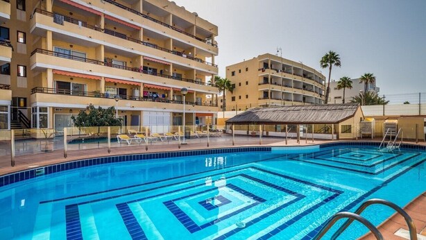 Une piscine aux carreaux bleus, dotée d’un parasol, entourée d’un immeuble à plusieurs étages avec des balcons, et un palmier en arrière-plan.