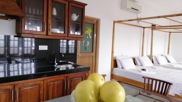 Una cocina con armarios de madera y una encimera negra, una cama con estructura de madera y ropa de cama blanca, y un cuenco con fruta amarilla sobre la mesa.