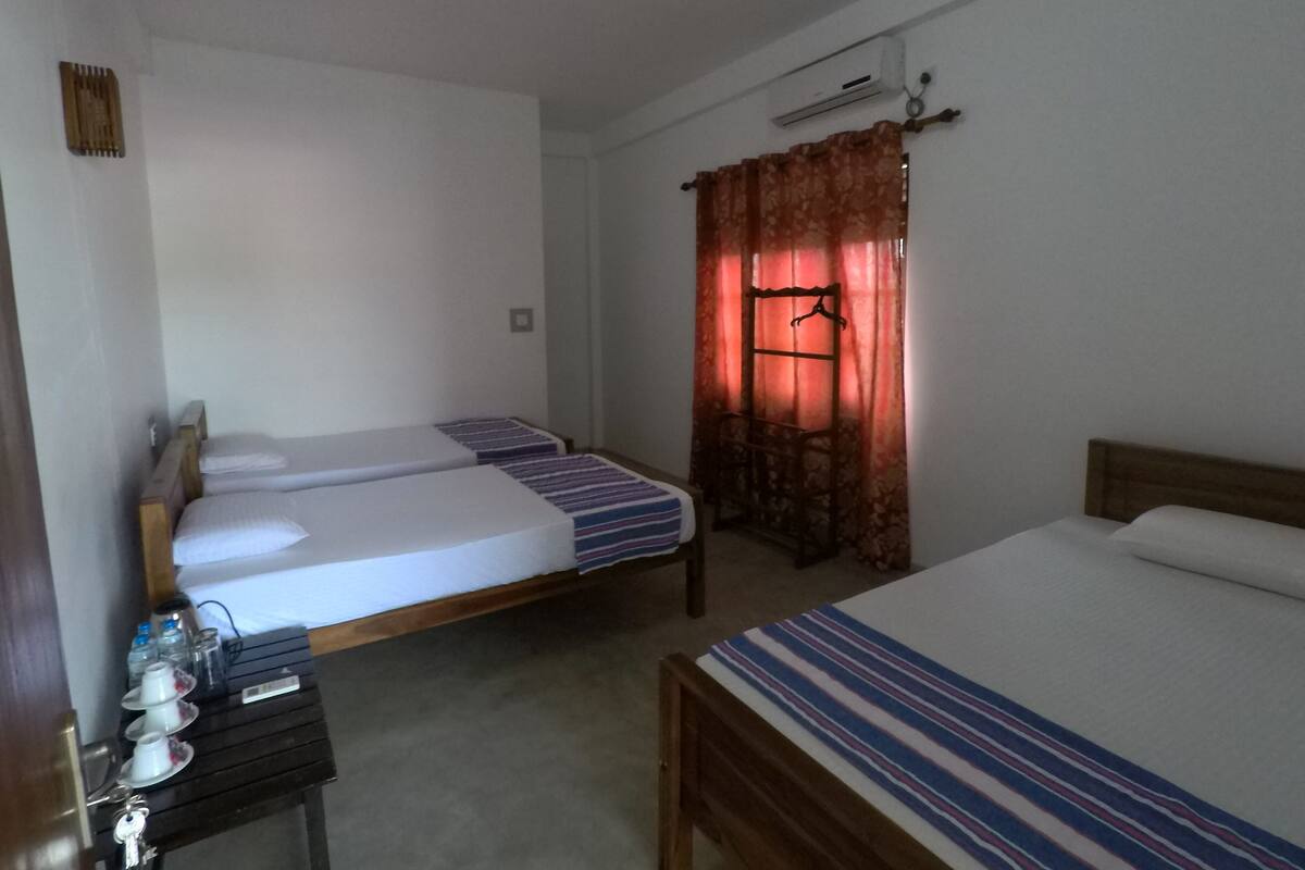 A hotel room with two single beds, a wooden headboard, a small table with toiletries, and a window with red curtains.