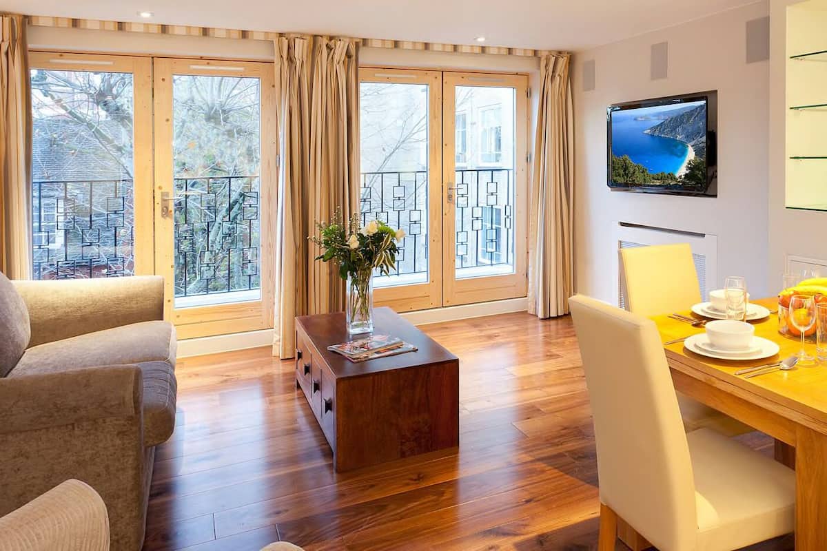 A modern living room with a wooden dining table set for two, a sofa, and a TV mounted on the wall.