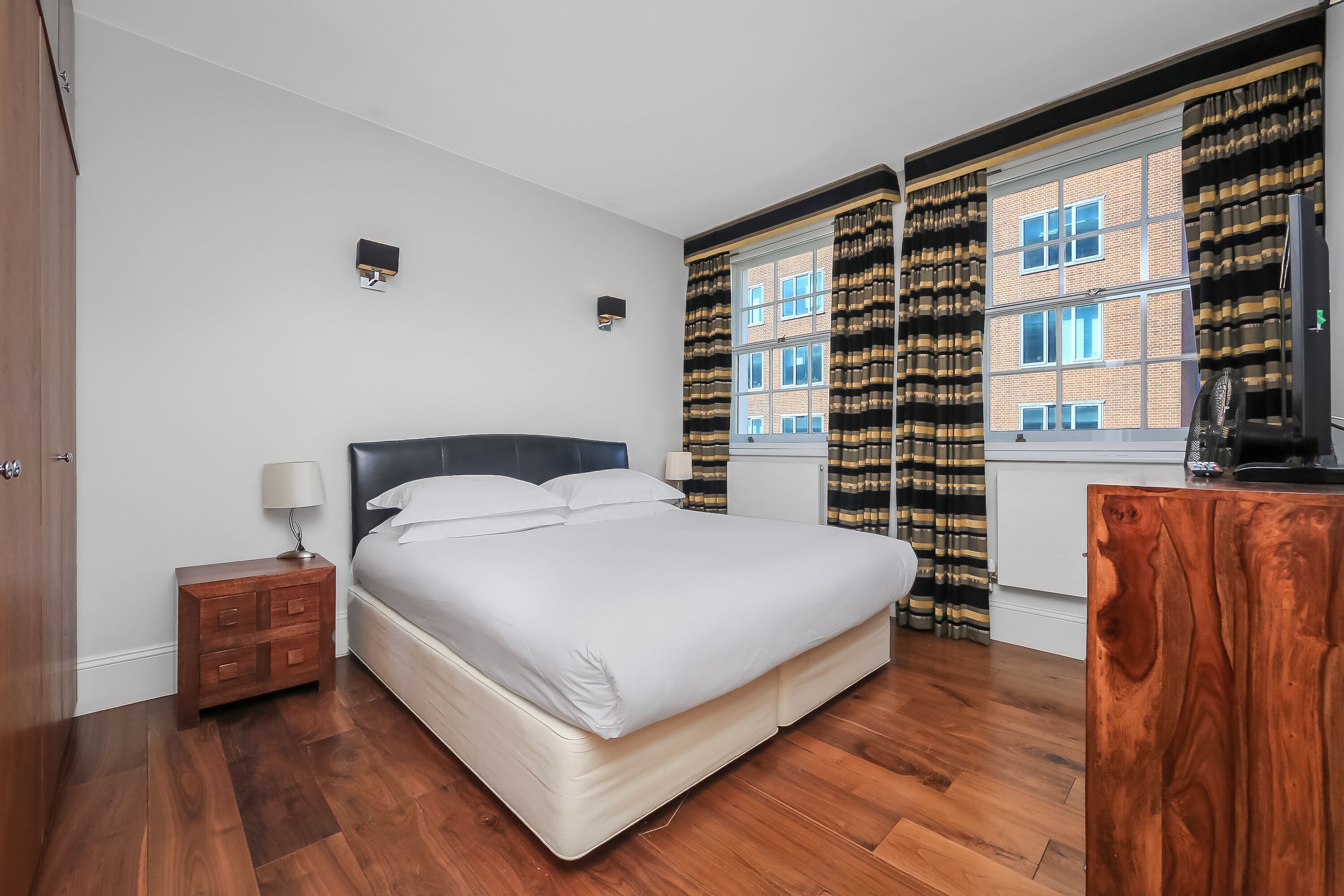 A neatly arranged bedroom with a large bed, a nightstand, a wooden dresser, and a view of a building through the window.