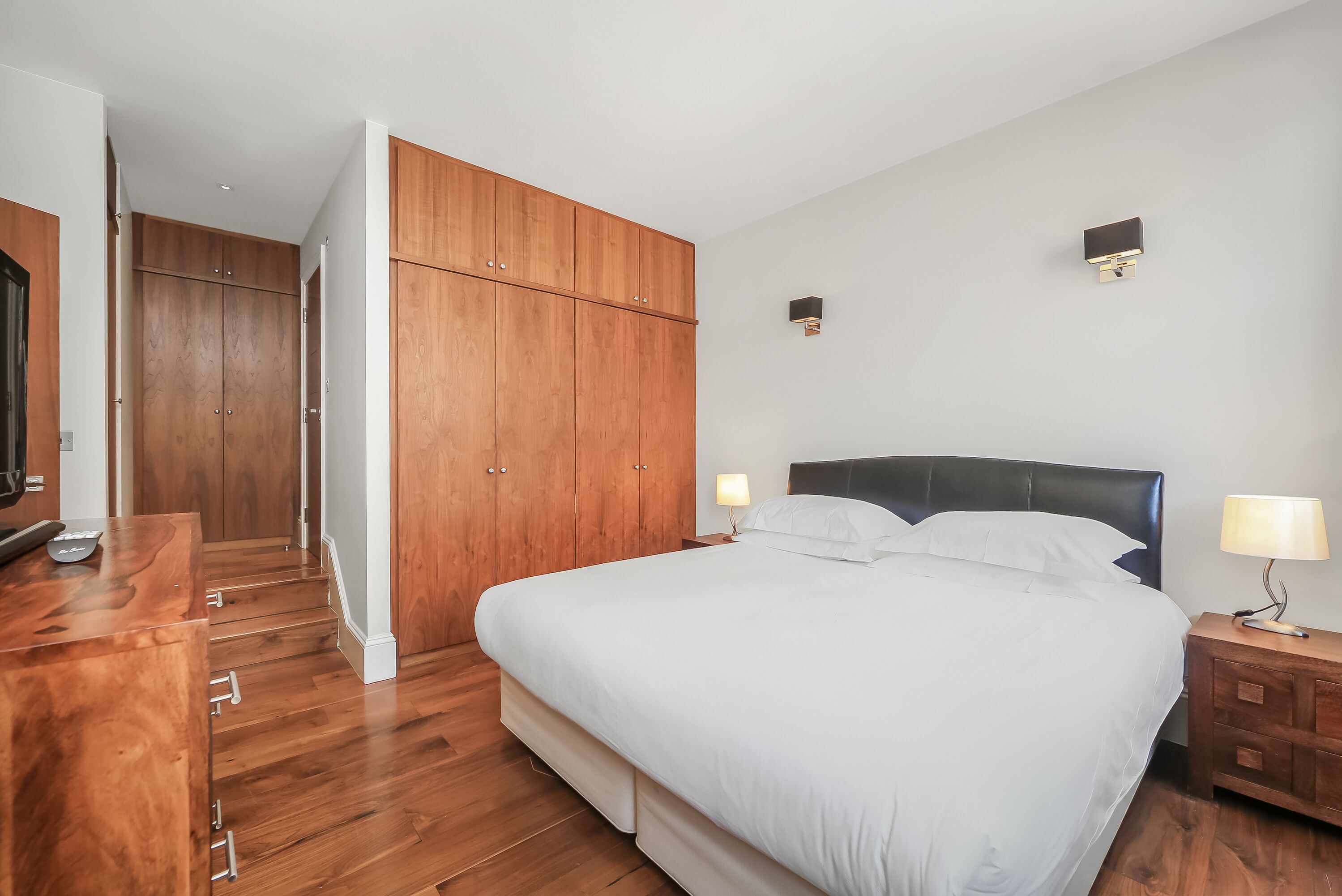 A bedroom with a large bed, wooden wardrobe, bedside tables with lamps, and a wooden desk.