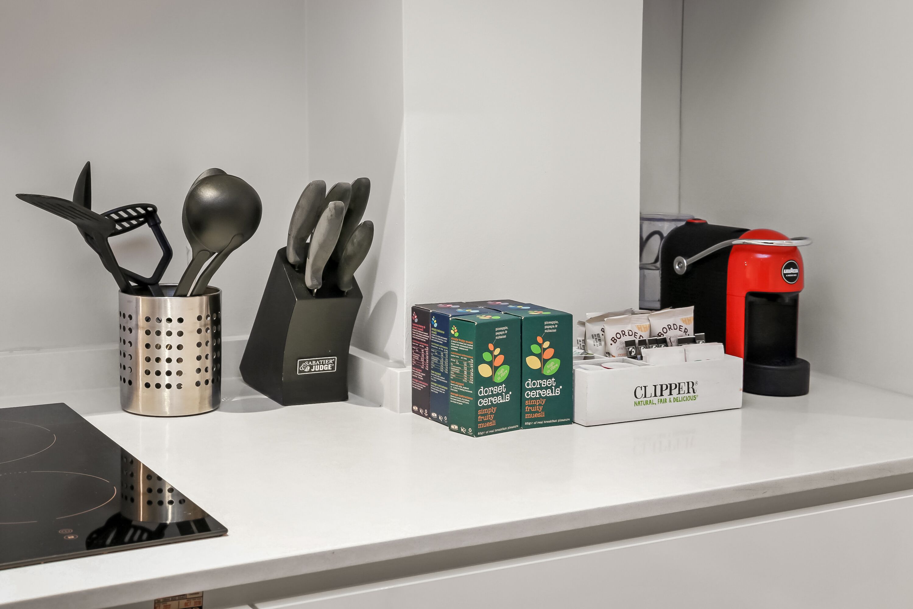 A kitchen counter with a colander, a spatula holder, a box of Dorset cereals, and a red electric kettle.
