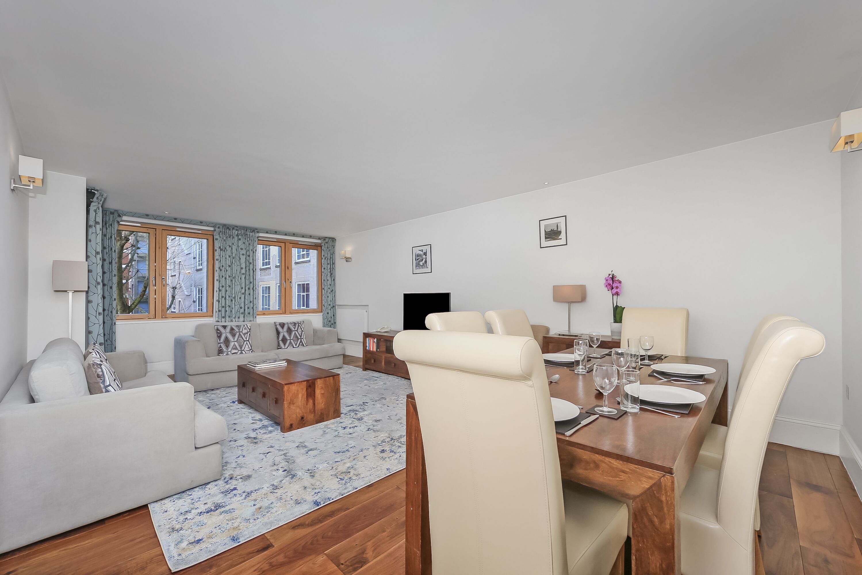 Una sala de estar moderna con una mesa de centro de madera, sofás beige, una mesa de comedor para cuatro personas y ventanas grandes con cortinas.