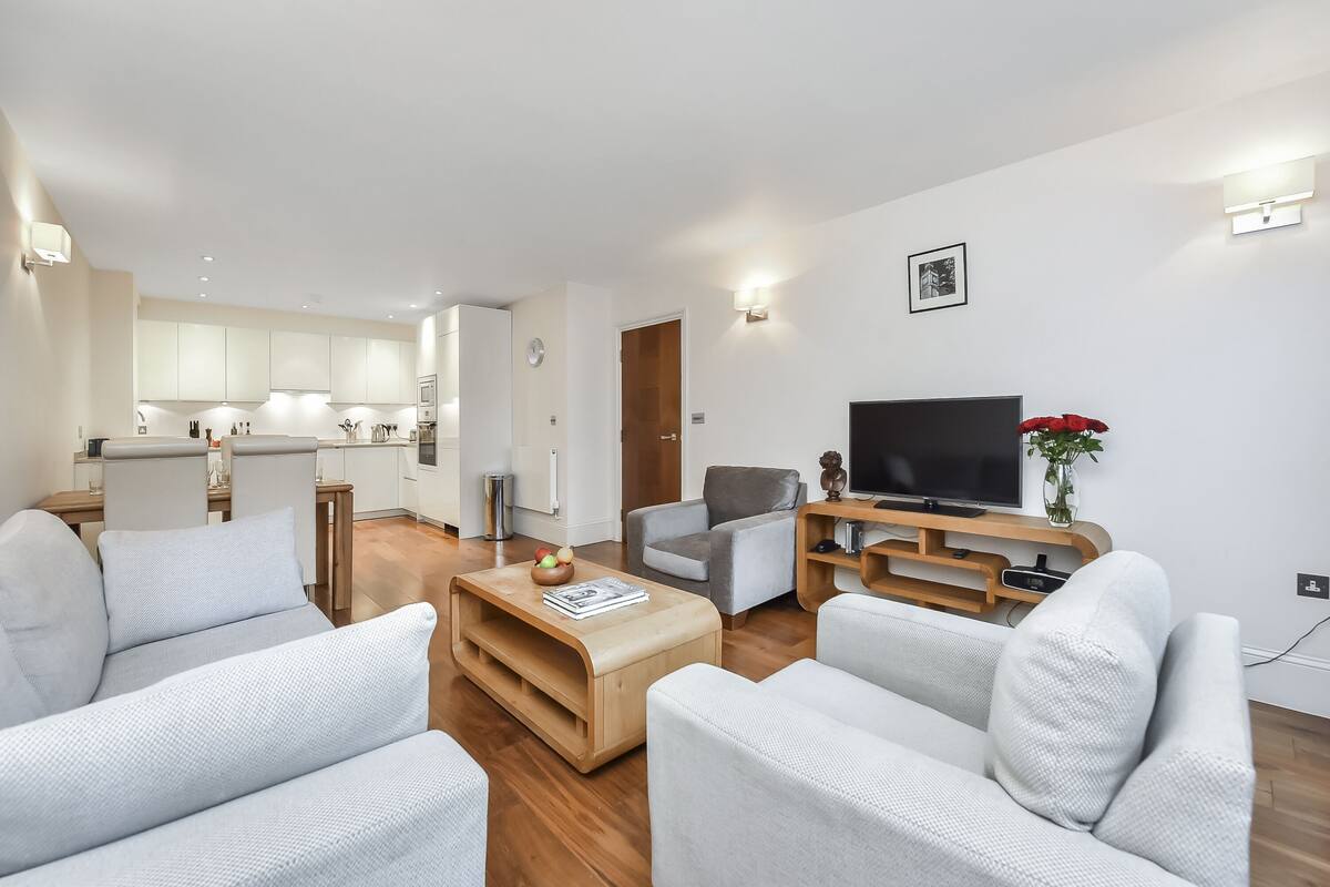A modern living room with a sofa, a coffee table, and a TV. There is a kitchen in the background with white cabinets and a wooden door.
