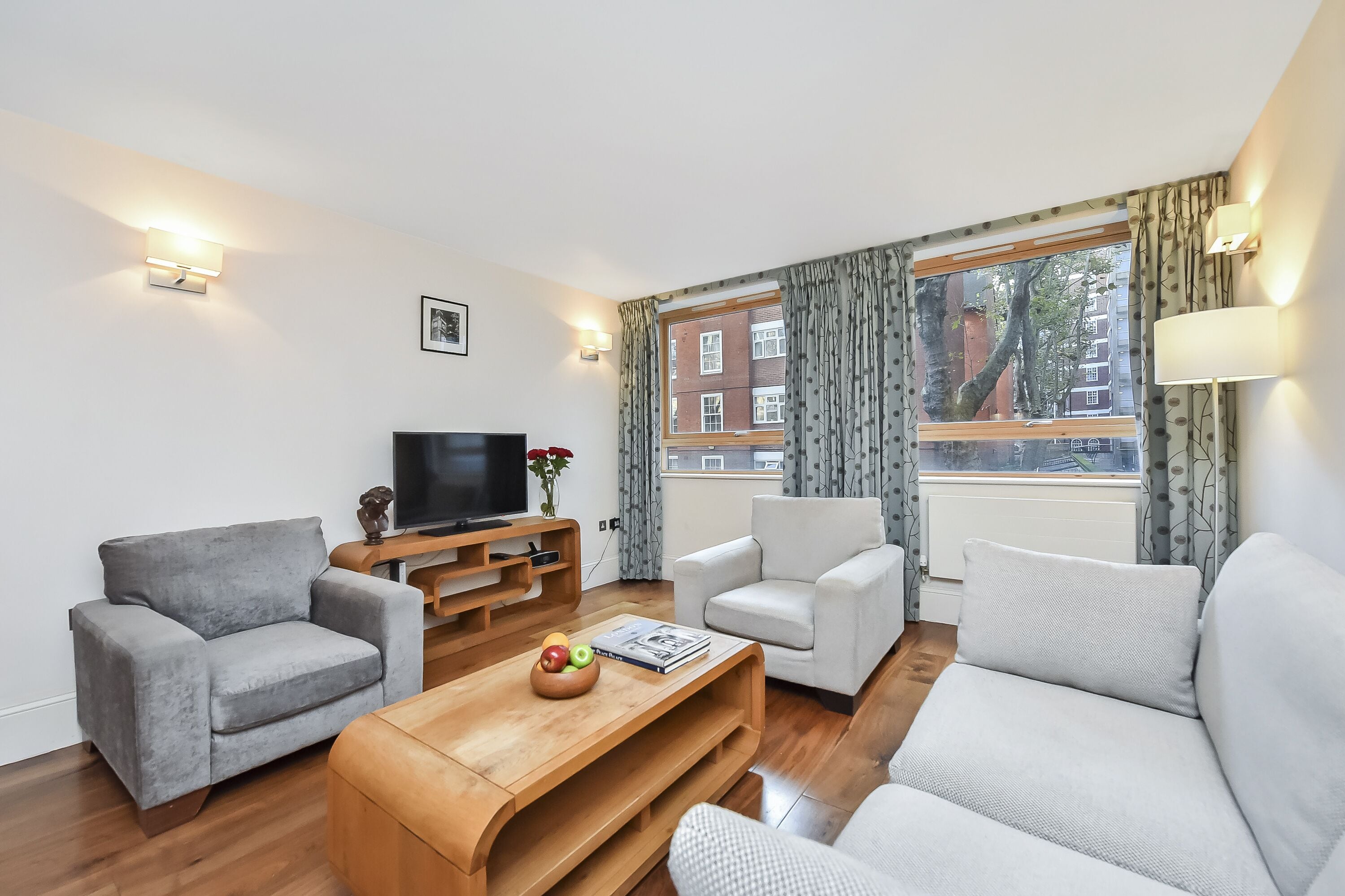 A modern living room with a grey sofa, a white armchair, a wooden coffee table with a fruit bowl, and a flat-screen TV on a wooden stand.