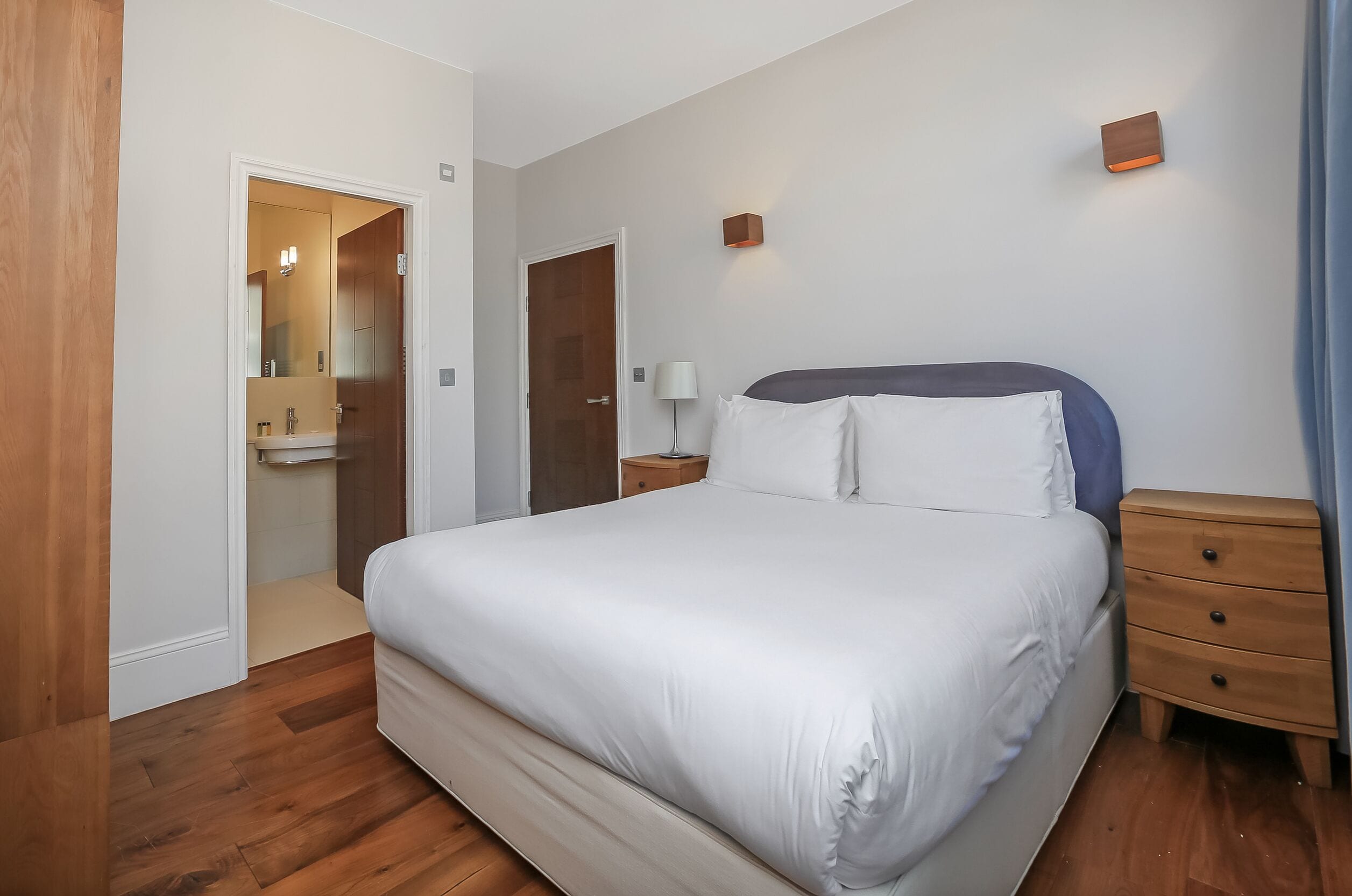 Quarto com uma cama grande, uma mesa de cabeceira de madeira, uma mesinha de apoio, uma luminária na parede e uma porta que dá para o banheiro.
