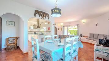 A dining area with a table set for six, white chairs, a chandelier, and a view of the kitchen and living area.