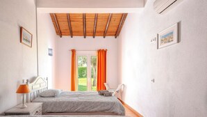 Ein Schlafzimmer mit zwei Betten, einem Fenster mit orangefarbenen Vorhängen, einer Holzbalkendecke und Blick ins Grüne.