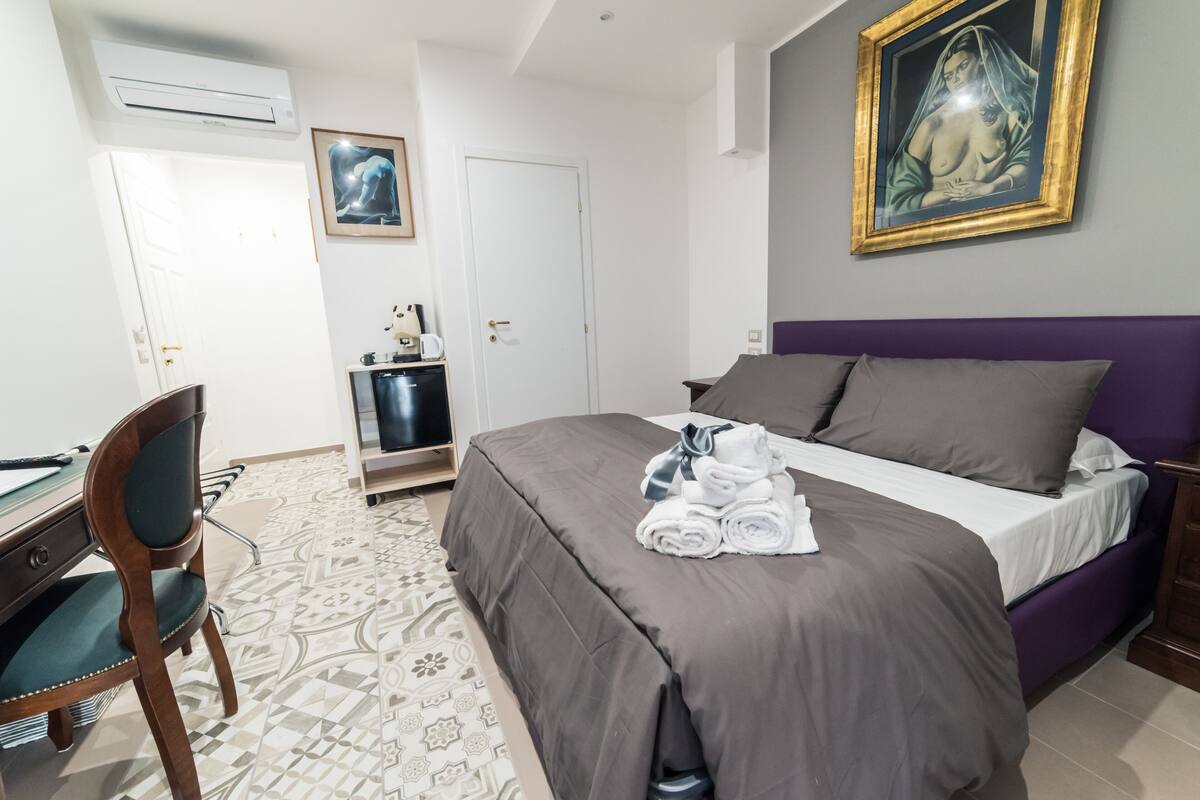 A hotel room with a bed, a desk with a chair, a bathroom, and a painting on the wall.