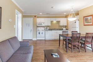 Una sala de estar con un sofá gris, una mesa de comedor de madera con sillas, una mesita de centro y una cocina al fondo con electrodomésticos blancos.