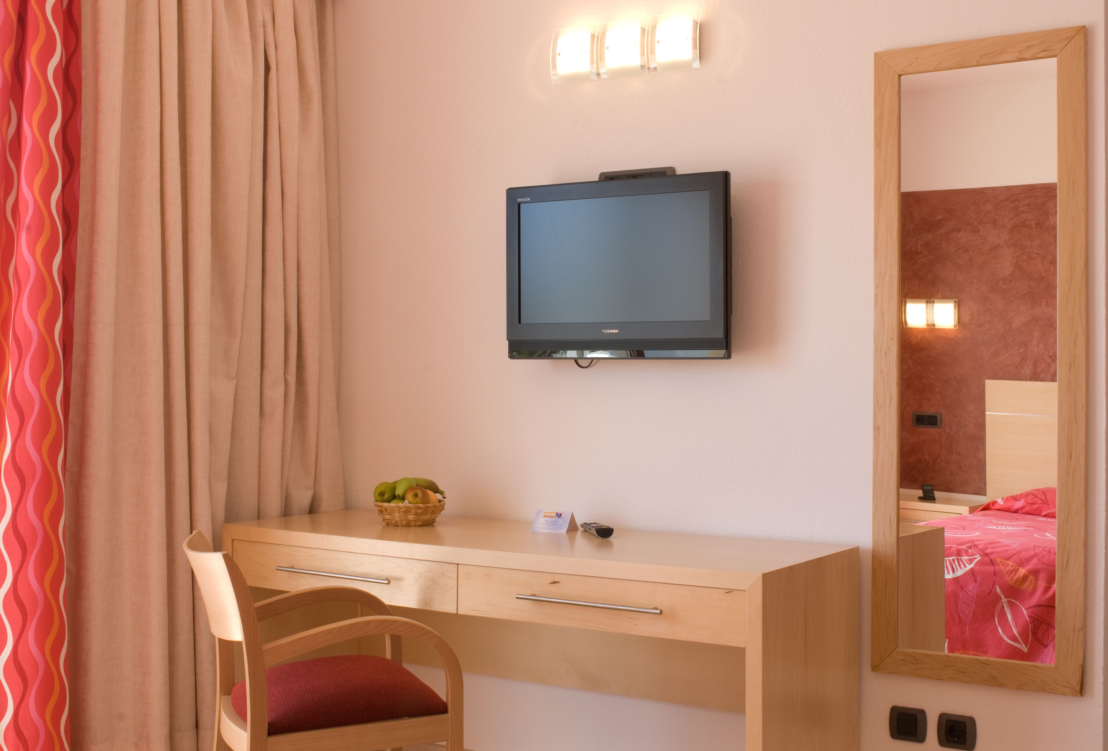 Un dormitorio con un escritorio de madera, televisión montada en la pared, un espejo y una cama con colcha roja.