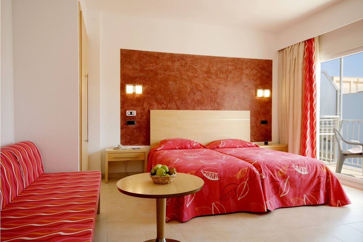 A hotel room with a bed, a small table with a fruit basket, a nightstand, and a view of the outside.