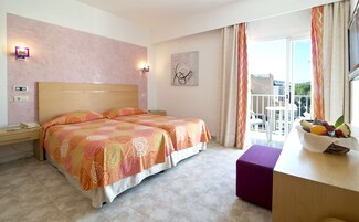 Habitación de hotel con dos camas, un escritorio con frutero, vista a la ciudad y balcón con una silla.