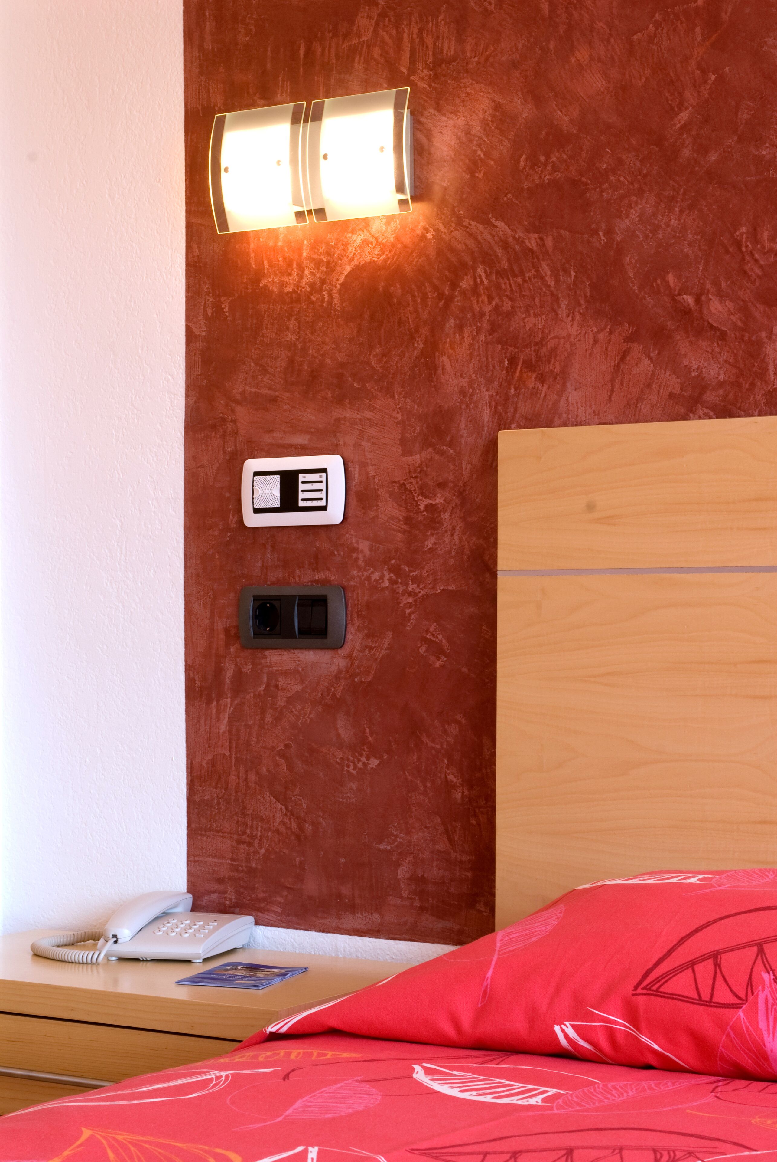 Quarto de hotel com cabeceira de madeira, mesa de cabeceira com telefone, abajur e travesseiro vermelho.