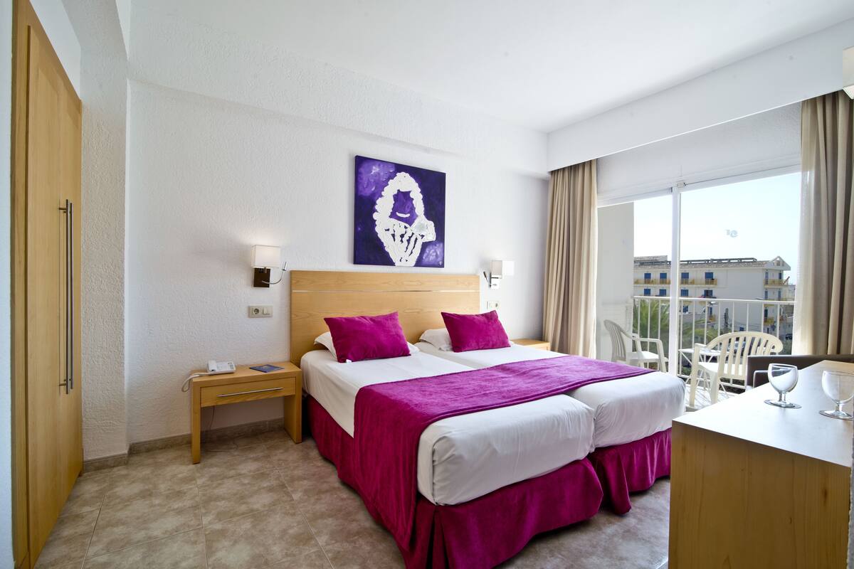 A hotel room with a large bed, two bedside tables, a desk with a chair, and a window with a view of buildings.