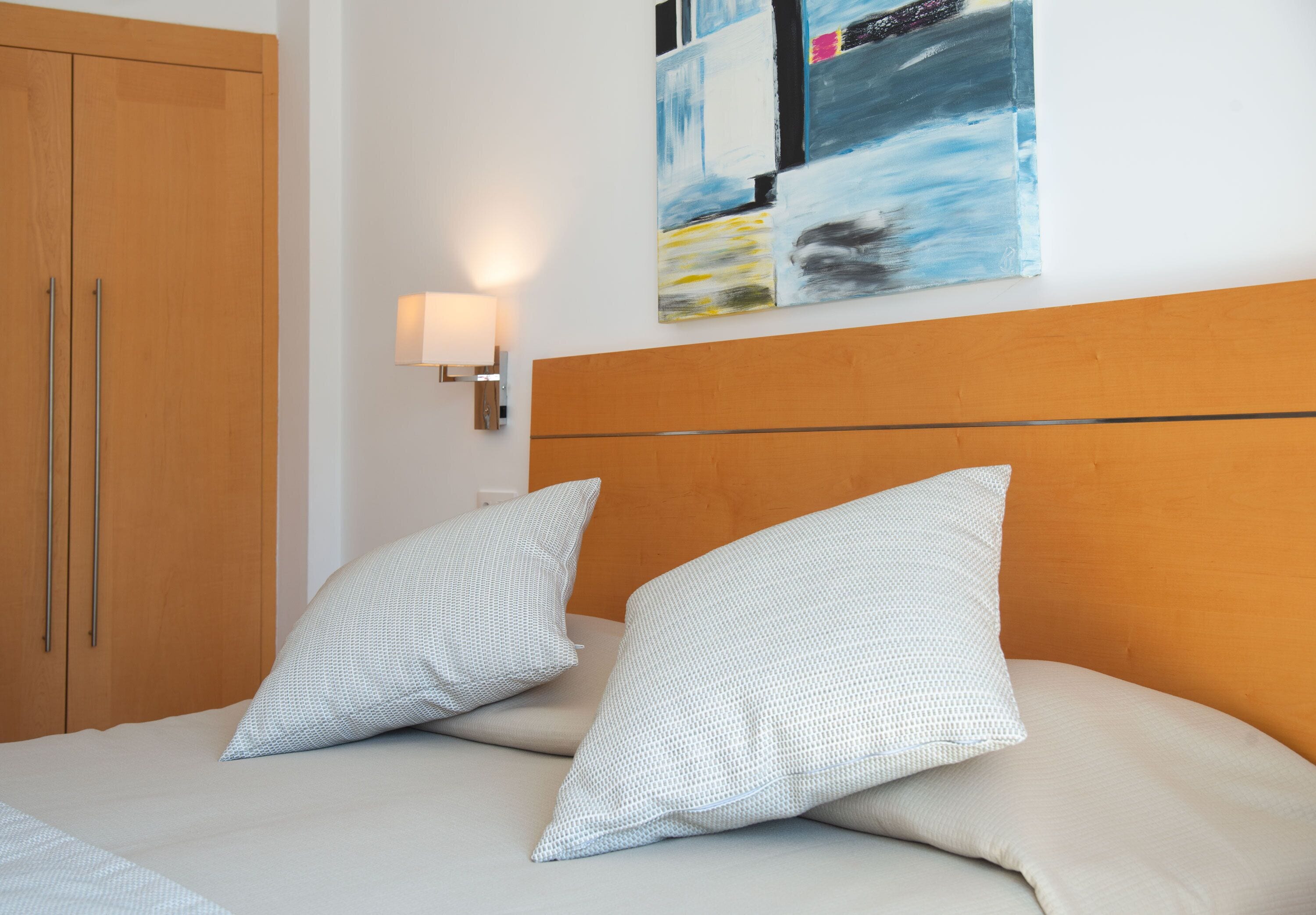 Un dormitorio con cama, almohadas, una mesita de noche, una lámpara de pared y un cuadro.