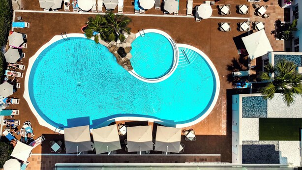 An aerial view of a hotel with a large, irregularly shaped swimming pool surrounded by lounge chairs and umbrellas. - Hotel Capricho (Capdepera)