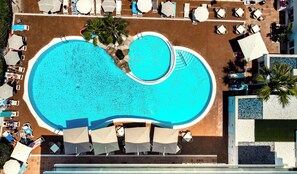 Vista aérea de un hotel con una piscina grande y de forma irregular, rodeada de sillones de descanso y sombrillas.