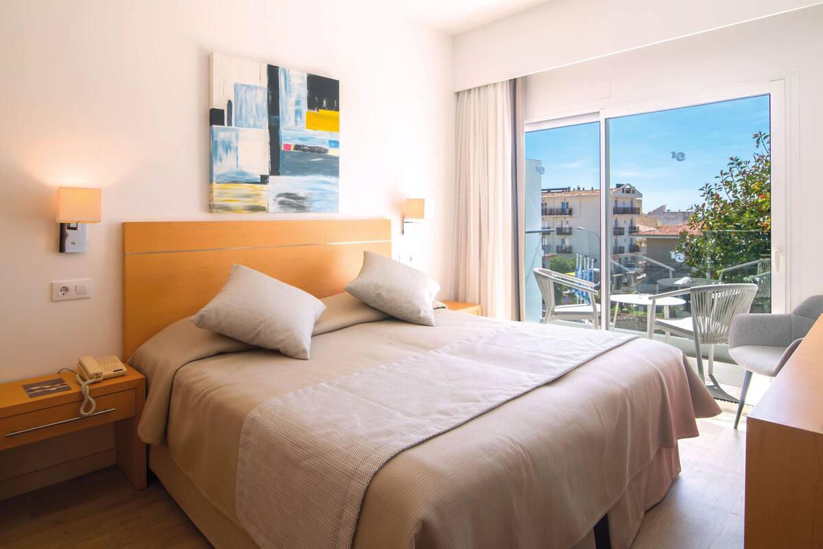 A hotel room with a large bed, a bedside telephone, a painting on the wall, and a balcony with a view of buildings.