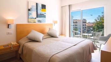 A hotel room with a large bed, a bedside telephone, a painting on the wall, and a balcony with a view of buildings.