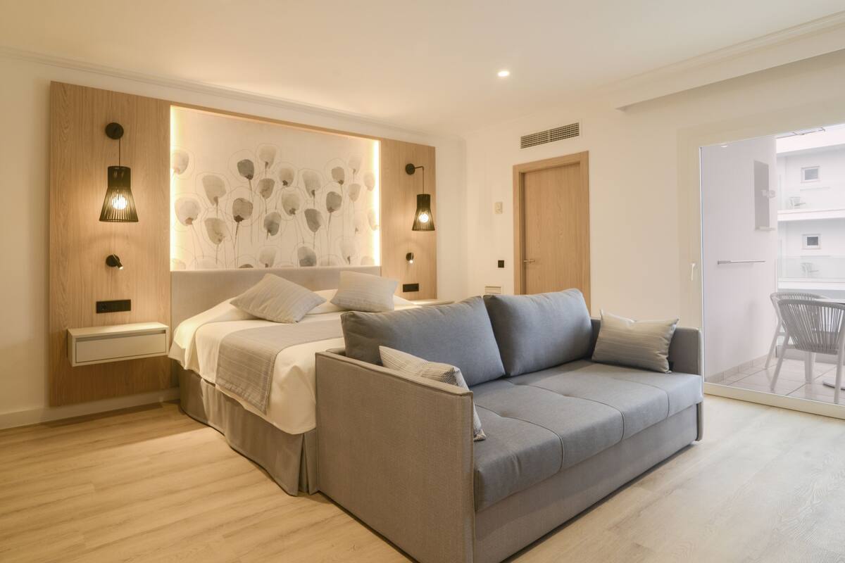 A modern bedroom with a grey sofa, a bed with white bedding, and a wall-mounted light fixture.