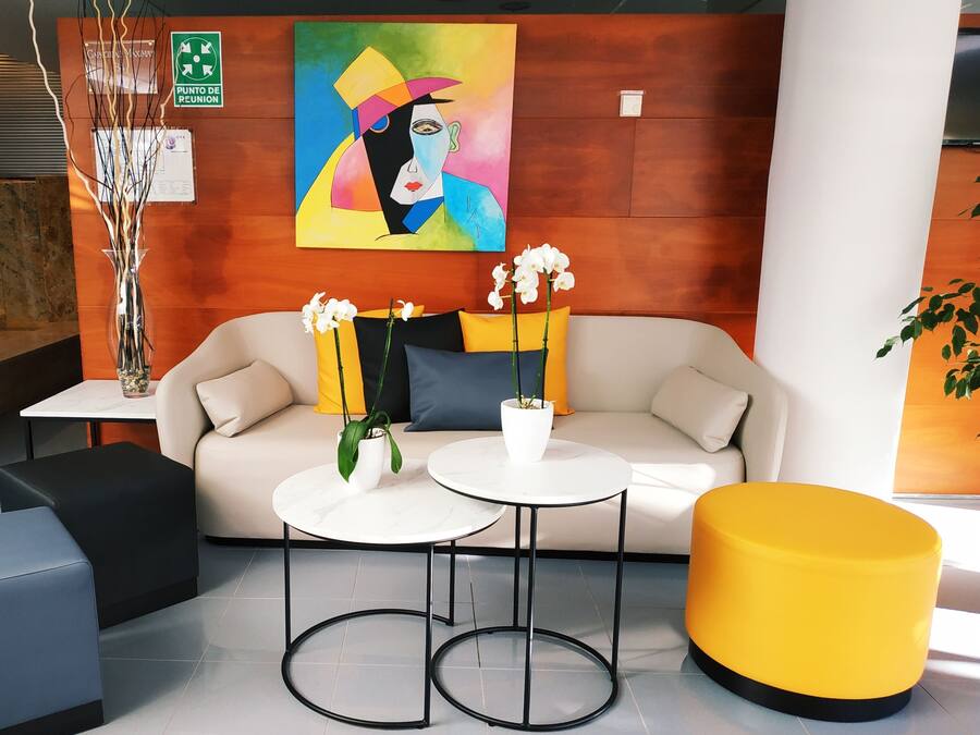 A modern lobby with a beige sofa, two white round tables, a yellow ottoman, and a colorful abstract painting on the wall.