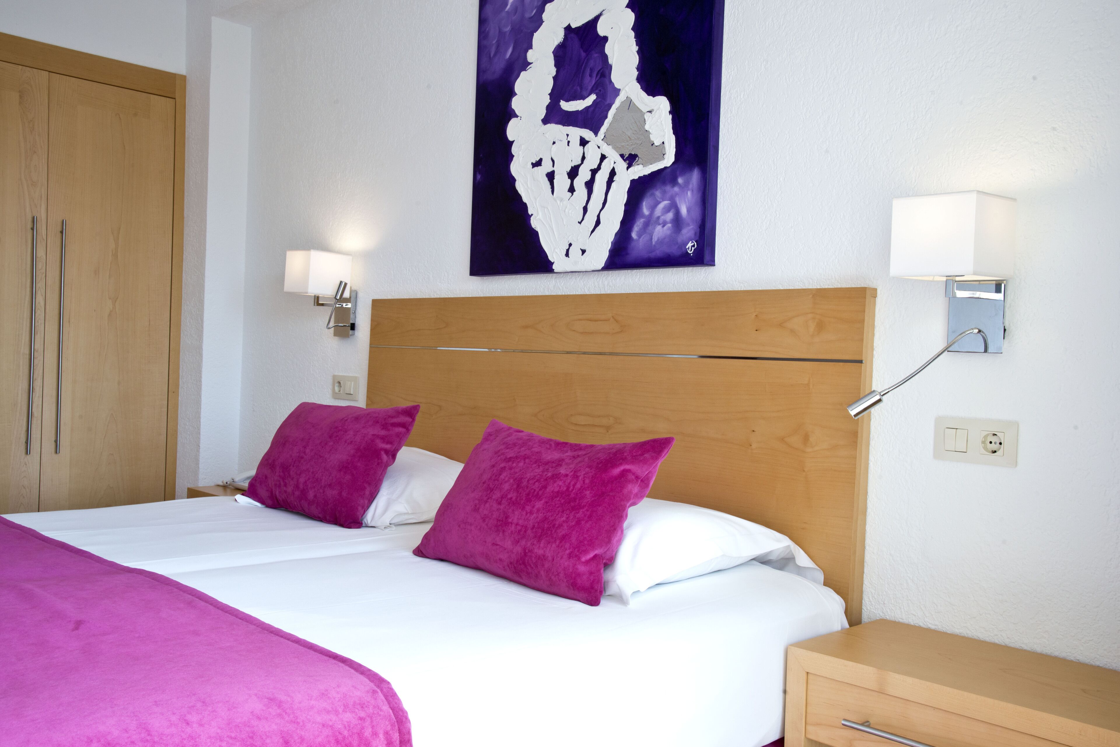 Une chambre d’hôtel avec une tête de lit en bois, deux oreillers violets, une couvre-lit blanche et un tableau au mur.