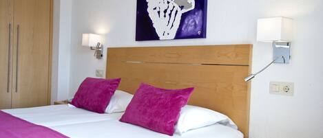 Une chambre d’hôtel avec une tête de lit en bois, deux oreillers violets, une couvre-lit blanche et un tableau au mur.
